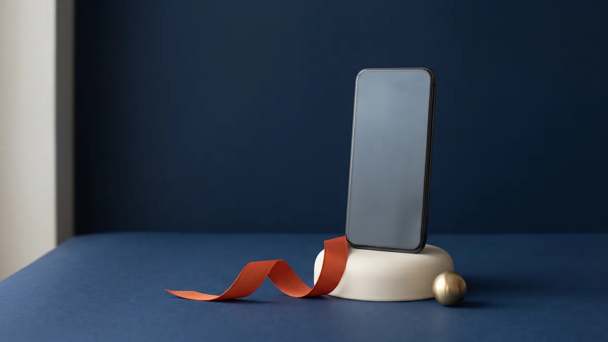 Smartphone standing on a display plinth with an orange ribbon, the cover image for iPhone is Kicking Arse